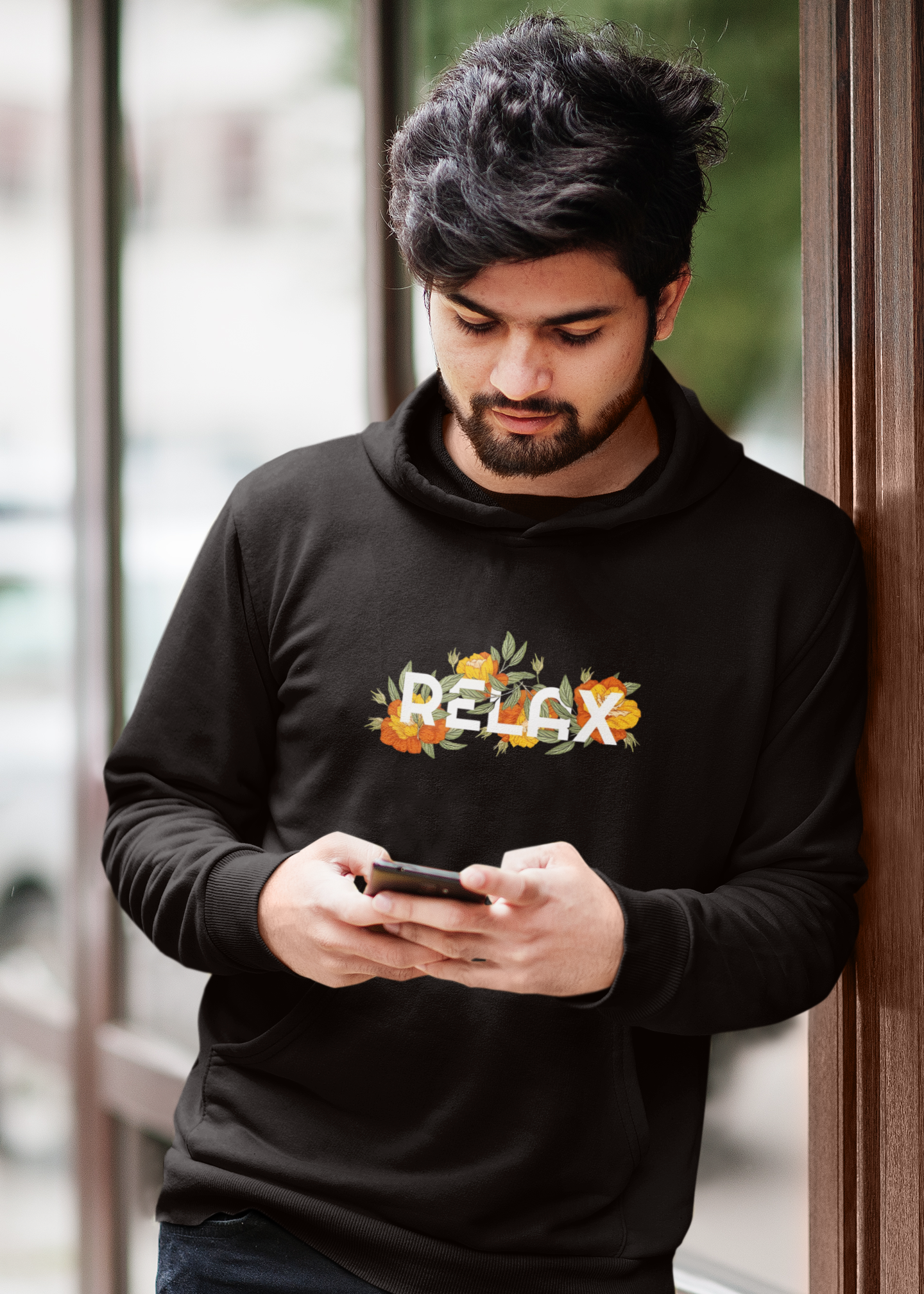 Man wearing a black hoodie with floral text design, leaning against a door.