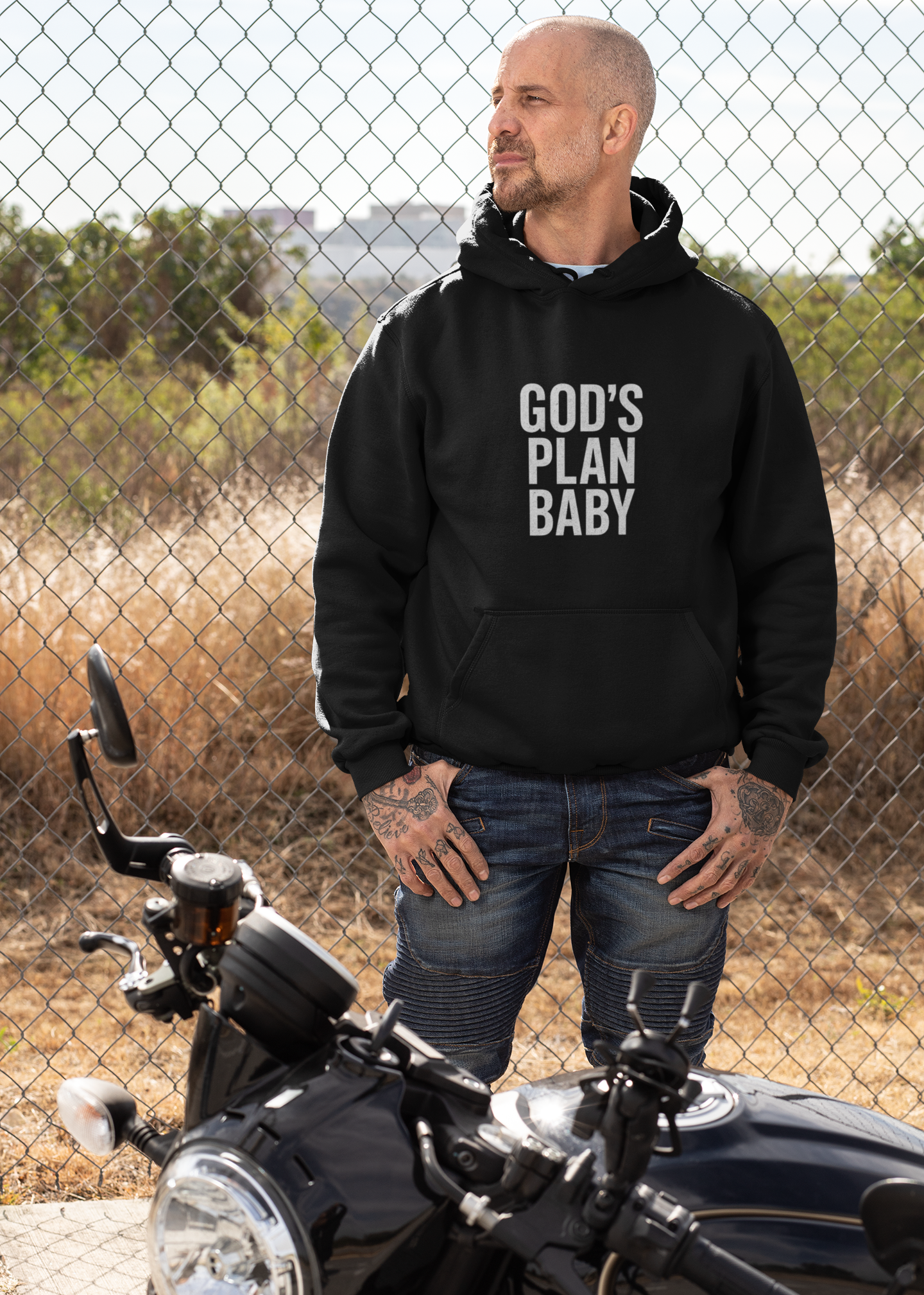 Man wearing a black hoodie with 'GOD'S PLAN BABY' text standing next to a motorcycle.